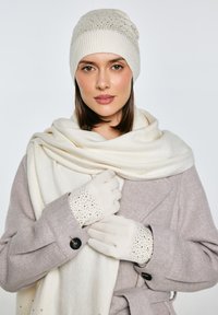 Béžová pletená čepice se zlatými akcenty, krémová šála a šedý kabát s rybí kostí vzorem. Krémové rukavice s ozdobnými detaily na prstech.