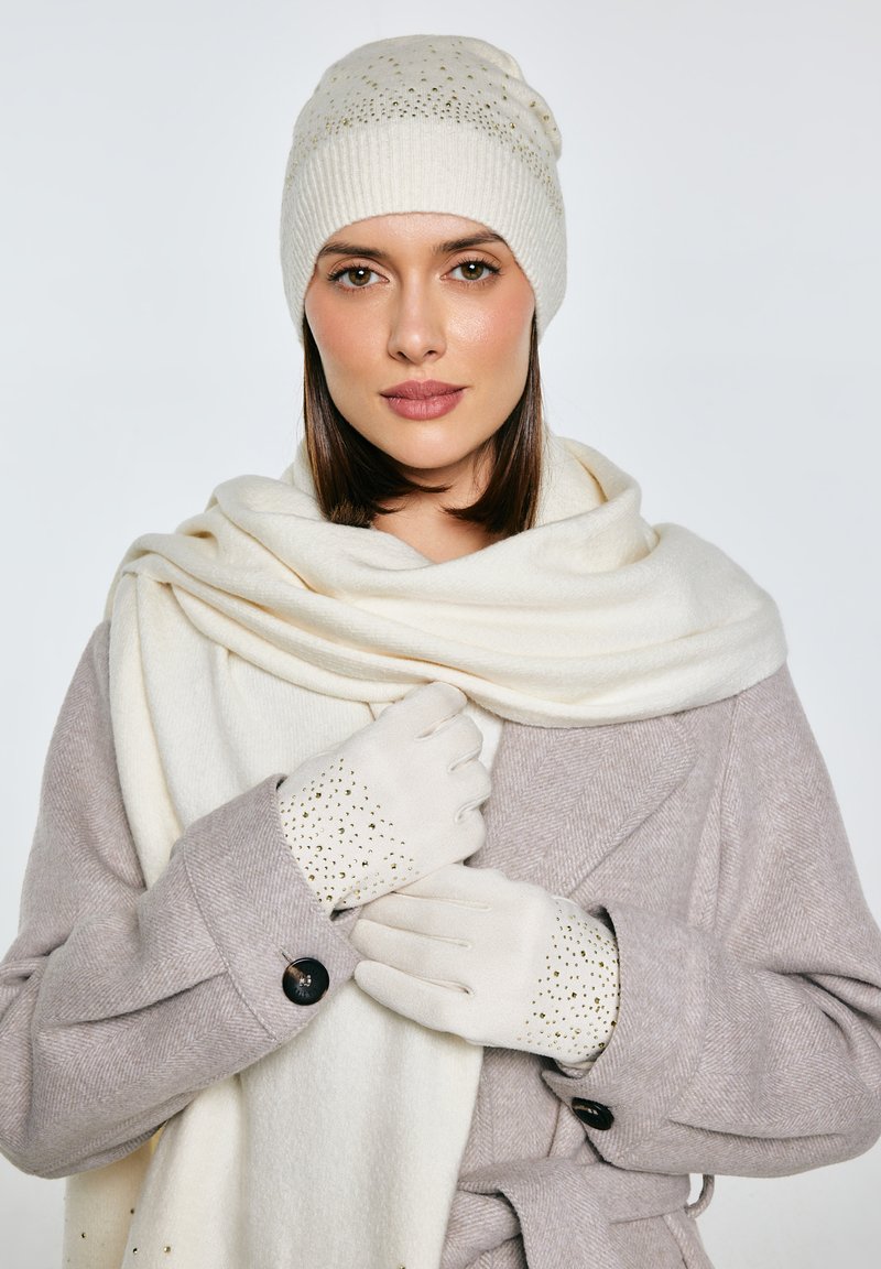 Béžová pletená čepice se zlatými akcenty, krémová šála a šedý kabát s rybí kostí vzorem. Krémové rukavice s ozdobnými detaily na prstech.