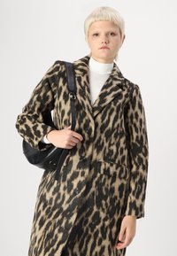 Abrigo de estampado de leopardo en beige y negro, con cuello de muesca, botones y bolsillos frontales. Acompañado de un bolso de hombro negro.