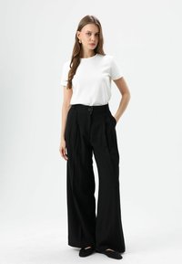 Weißes T-Shirt mit kurzen Ärmeln, kombiniert mit schwarzen, hoch taillierten, weit geschnittenen Hosen mit Knopfverschluss und Seitentaschen. Schwarze Ballerinas runden das Outfit ab.