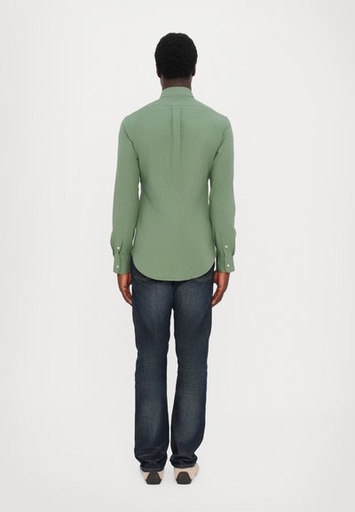 Chemise à manches longues verte avec un col, pli au dos et poignets boutonnés, associée à un jean en denim foncé et des chaussures beiges. Fond uni.