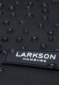 Tissu noir avec des gouttes d'eau perlant à la surface et une étiquette "Larkson Hamburg" attachée.