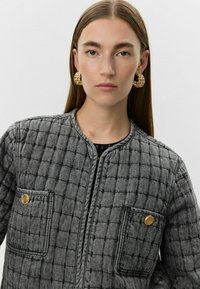 Chaqueta acolchada gris con cuello redondo, dos bolsillos frontales, botones dorados y una superficie texturizada. Acentos y detalles de costura en negro.