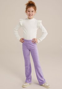 Bluză albă cu mâneci lungi și detalii cu volane la umeri, purtată împreună cu pantaloni din catifea ușoară de culoare mov deschis, evazați. Modelul stă cu mâinile pe șolduri, zâmbind.