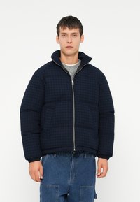 Marineblå dynejakke med rutemønster, høy krage, frontglidelås og to sidelommer, kombinert med en grå genser under.