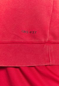 Gros plan sur un tissu rouge avec le texte imprimé "DRI-FIT" et des coutures visibles le long de l'ourlet, montrant un tricot texturé et des couches de matériau lisse.