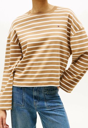 Person trägt ein beige-weiß gestreiftes Langarmshirt mit kleinem Logo auf der Brust, kombiniert mit blauen Jeans.