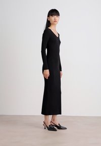 Woman standing sideways wearing a long black fitted dress and black kitten-heel slingback shoes against a plain white background.