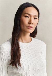 Femme asiatique aux longs cheveux foncés portant un pull blanc à torsades sur un fond neutre uni, regardant calmement la caméra.