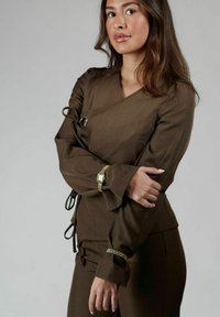 Top marrone a maniche lunghe con scollo a V e maniche arricciate, dotato di hardware decorativo e finitura testurizzata, indossato con pantaloni coordinati.