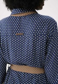 Personne portant une chemise bleue à motifs et une ceinture marron avec des motifs géométriques, montrant son dos et ses épaules, avec des cheveux bouclés partiellement visibles.