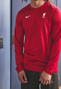 Rotes Langarmshirt aus atmungsaktivem Stoff, mit einem weißen Nike-Logo und dem Wappen des Liverpool FC auf der linken Brust. Glatte Textur.