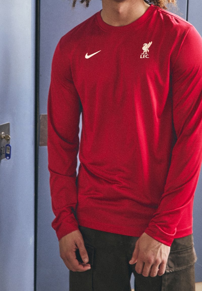 Rotes Langarmshirt aus atmungsaktivem Stoff, mit einem weißen Nike-Logo und dem Wappen des Liverpool FC auf der linken Brust. Glatte Textur.