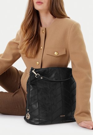 Mujer con abrigo color camello con botones dorados sostiene un gran bolso de cuero negro acolchado con adorno metálico y asa para el hombro.