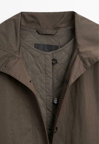 Brązowa pikowana kurtka z miękkiego materiału zewnętrznego, wyposażona w stójkę, ukośne przeszycia na podszewce i dwa czarne guziki.