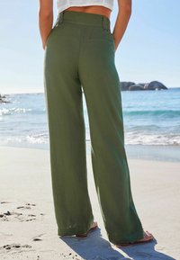 Pantalons oversized en lin vert avec taille haute, tissu texturé et poche arrière, sur fond de plage avec de l'eau et des rochers.