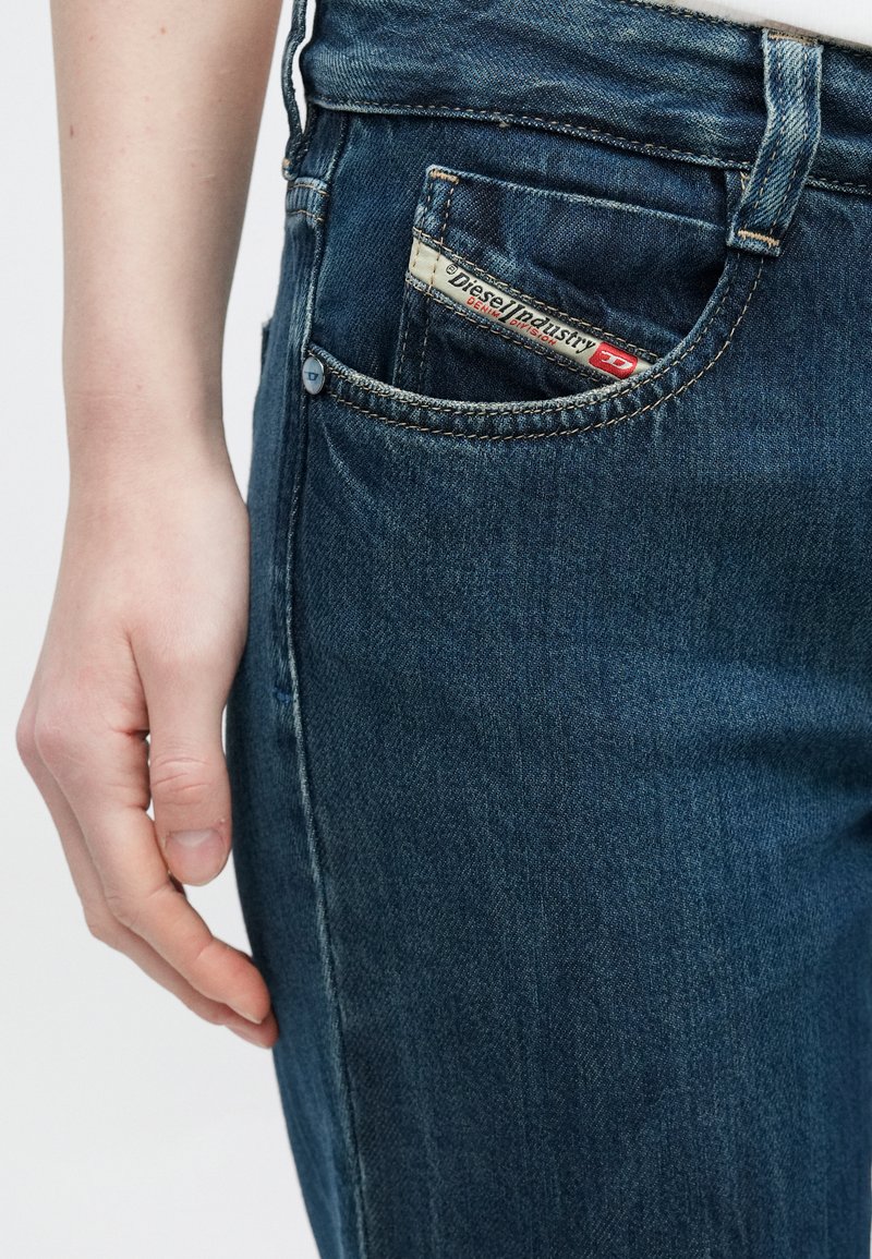 Close-up of a person’s hand resting beside dark blue Diesel jeans with a branded label visible on the small front pocket.
