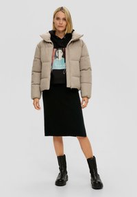 Beige puffer jack met een hoge kraag, gedragen over een zwarte hoodie met een grafische print, gecombineerd met een zwarte geribde rok en chunky zwarte laarzen.