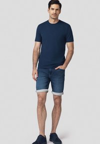 Granatowa bawełniana koszulka, w połączeniu z jasnoniebieskimi szortami dżinsowymi z podwiniętymi mankietami i granatowymi sneakersami, na neutralnym tle.