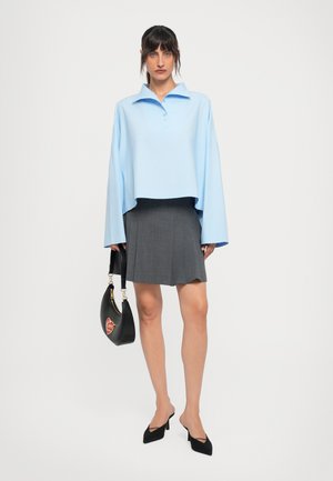 Woman wearing a light blue oversized collared shirt, gray pleated skirt, black heels, and holding a black handbag with a red heart patch.