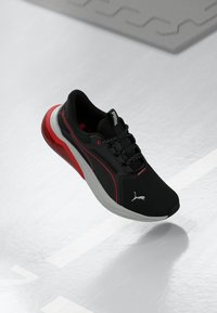 Svart atletisk sko med röda accenter, mesh-överdel och snördesign. Har en dämpad sula och Puma-logotyp på sidan och tungan.