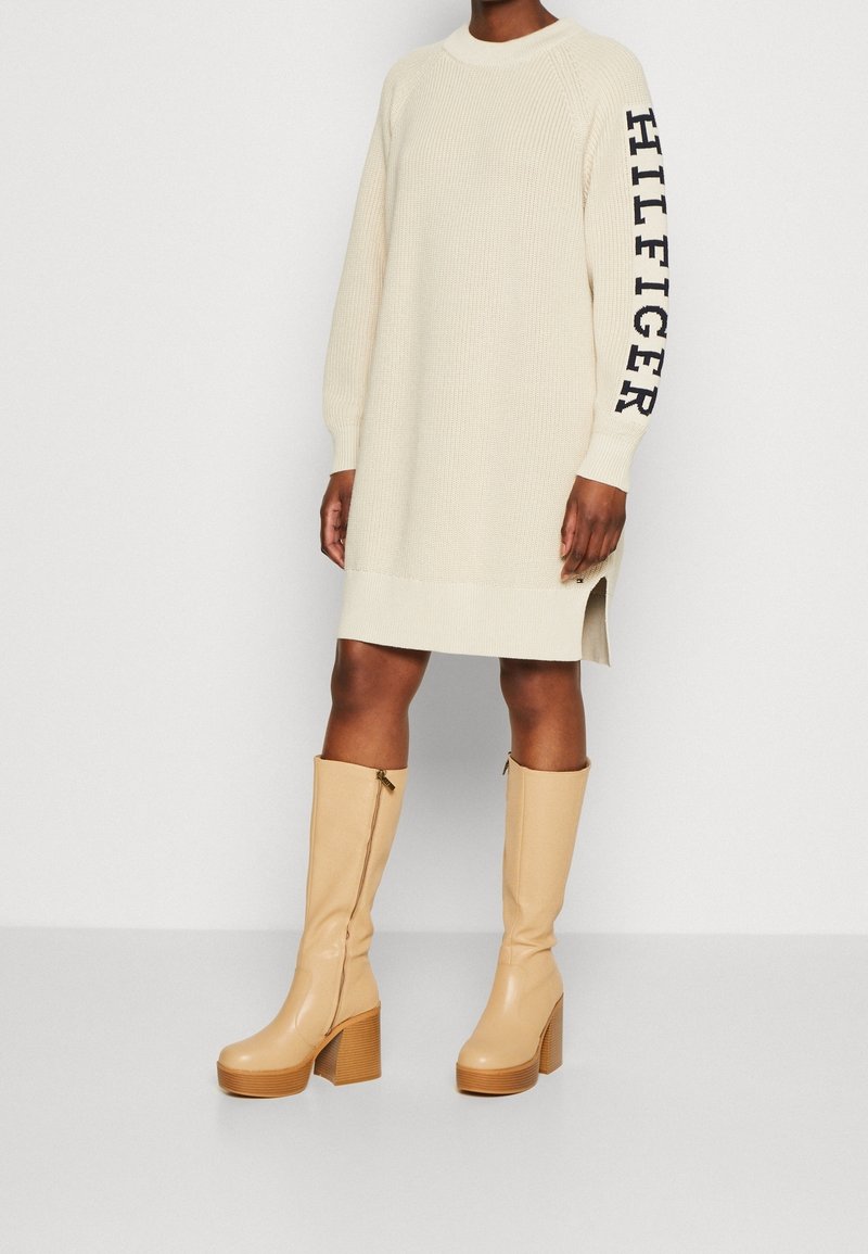 Abito lavorato a maglia crema con spacchi laterali, caratterizzato dal testo "HILFIGER" sulla manica. Abbinato a stivali color beige al ginocchio con tacco.
