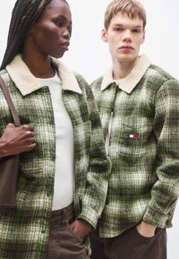 Groene geruite overhemden met een crèmekleurige sherpa-kraag en een gestructureerde stof, gedragen door twee personen. Eén houdt een bruine tas vast, wat een casual stijl toont.