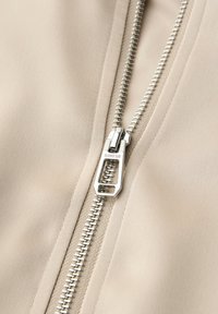 Gros plan sur une fermeture éclair en métal argenté partiellement fermée sur un tissu beige, avec l'inscription "STREET ONE" gravée sur le tirette de la fermeture éclair.