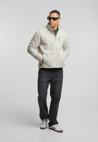 Fleecejakke i off-white med en lynlåslomme, høj krave og struktureret stof. Bæres med mørke jeans og sneakers.