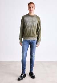 Sudadera de cuello redondo en verde oliva con un acabado texturizado, combinada con jeans ajustados de color azul claro y zapatos de cuero negros.