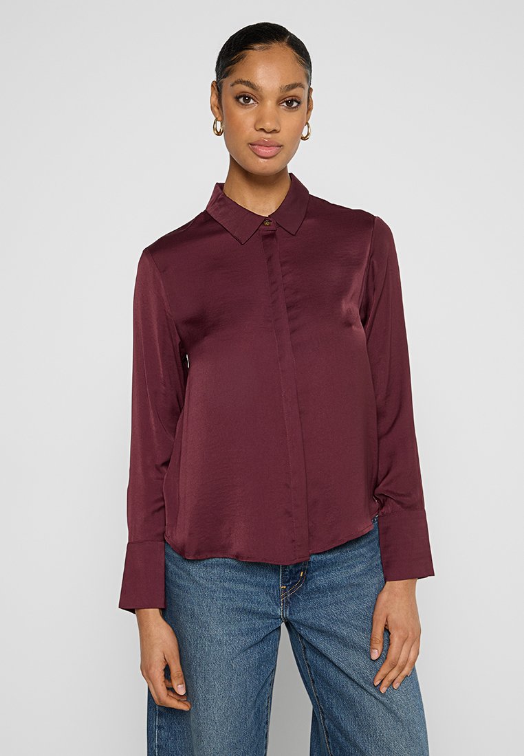 Burgunderrote Satinbluse mit Kragen, Knopfleiste vorne und langen Ärmeln. Glatte Textur und leichter Glanz. Kombiniert mit blauen Jeans.