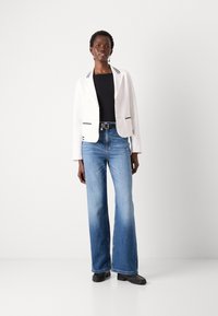 Blazer blanc avec bord noir, haut noir et jeans large bleu clair. Chaussures noires avec détail texturé. Se tenant contre un fond uni.