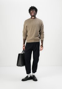 Maglione a maglia marrone, pantaloni neri a sigaretta, scarpe nere e calzini bianchi. Tiene una borsa a tracolla in pelle nera. Sfondo neutro.
