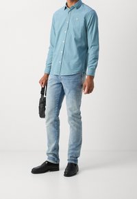 Camicia azzurro chiaro a maniche lunghe con bottoni, jeans blu sbiaditi, mocassini neri e borsa a tracolla nera. Logo sottile sulla camicia.