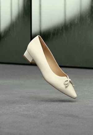 Weißer flacher Lederschuh mit spitzer Zehe, kleinem Blockabsatz und einer dekorativen Schleife vorne, auf einer grauen Oberfläche präsentiert.