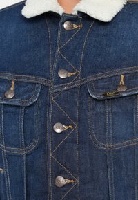 Giacca in denim blu scuro con un colletto in shearling bianco, bottoni metallici e dettagli di cucitura. Due tasche sul petto con accenti di marca.