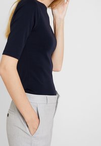 Kvinna i en svart figurnära kortärmad topp och ljusgrå skräddarsydda byxor, med en hand i fickan och håller ett vitt föremål nära sitt ansikte.