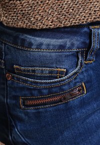 Vaqueros de mezclilla azules con un bolsillo trasero que presenta un detalle marrón con cremallera, costuras amarillas en contraste y una parte visible de un top de punto marrón texturizado.