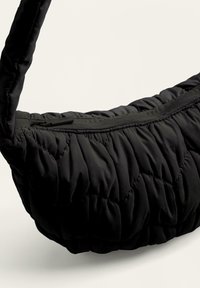 Svart quiltad handväska med en mjuk, puffig textur, med en bred rem och en dragkedjestängning på toppen. Materialet verkar slätt och lätt.