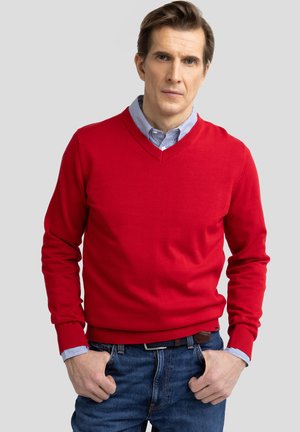 Suéter rojo de cuello en V hecho de material de punto fino, con puños y dobladillo de canalé. Llevado sobre una camisa de cuello azul claro con rayas.
