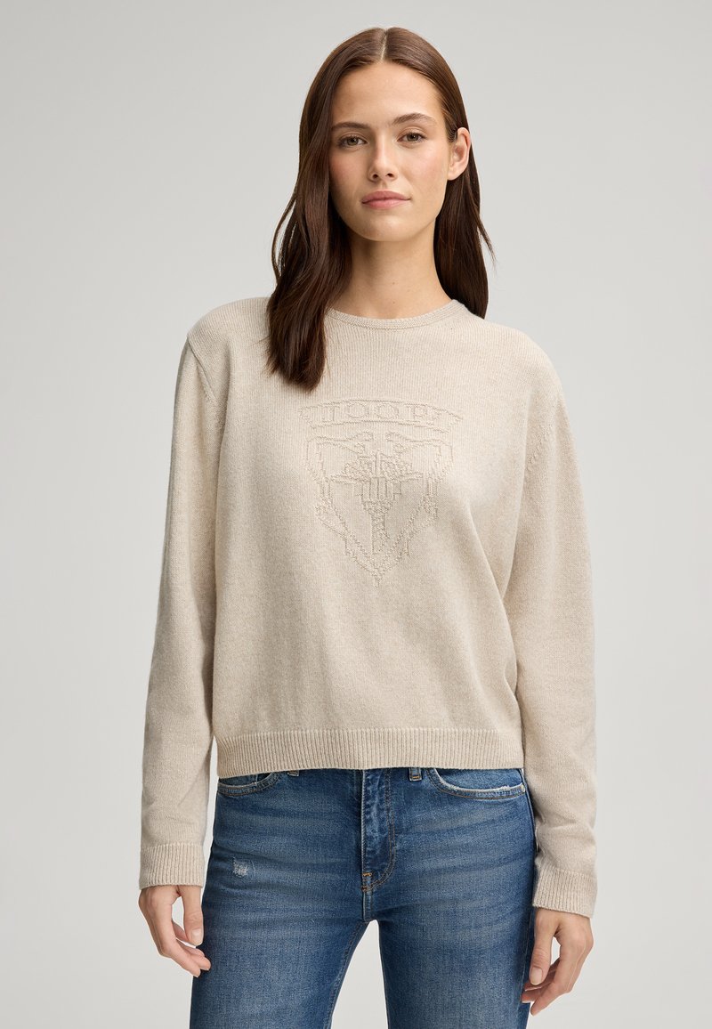 Maglione beige a girocollo con orlo e polsini a costine, con un sottile design del logo testurizzato sul davanti. Indossato con jeans blu.