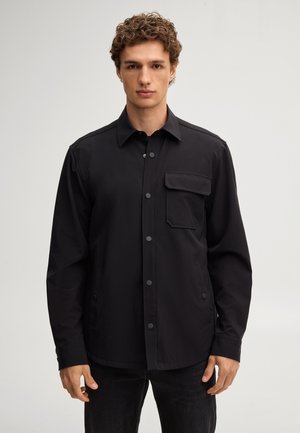 Jonge man die een zwart jasje met knopen draagt met een klepzak op de linkerborst, staand tegen een effen lichte achtergrond.