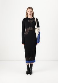 Lång svart stickad klänning med öppen meshdetaljering, med blå och vita accenter vid ärmarna och fållen. Modellen bär svarta klackboots.