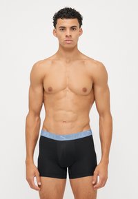 Fekete boxeralsó kék Puma logós derékkal, testhezálló fazon, sima textúra és közepes hosszúságú vágás. A modell karjait lazán tartja.