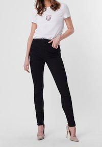 Svarta skinny jeans i kombination med en vit t-shirt med ett blommig kruka-mönster i rosa, bärs med nude högklackade skor. Slät tygtextur.