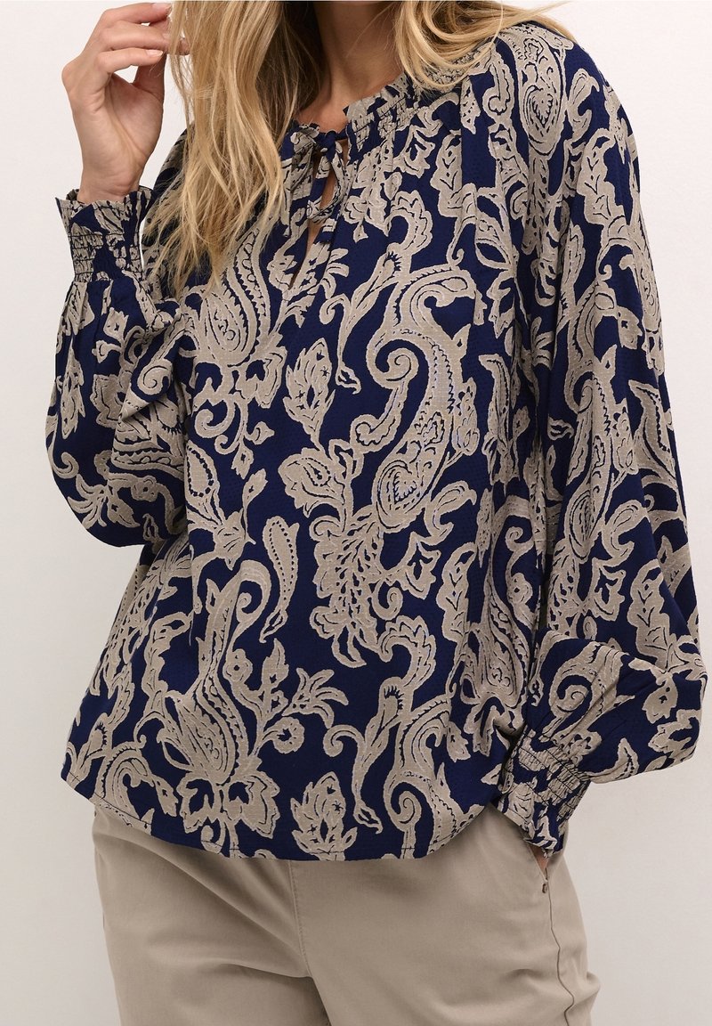 Femme portant un chemisier bleu marine avec des motifs floraux et cachemire beiges complexes et un pantalon beige, une main dans la poche et l'autre touchant ses cheveux.