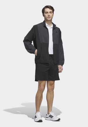 Fiatal férfi áll, fekete cipzáras dzsekit, fehér galléros pólót, fekete rövidnadrágot és fekete csíkos fehér tornacipőt visel, fehér háttér előtt.