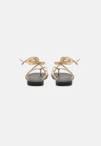 Sandales plates avec semelles noires, brides en fil doré formant un nœud à la cheville ; design minimaliste avec texture lisse et accents métalliques.
