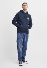 Mørkeblå hettegenser med frontlomme og grafisk detalj på brystet, kombinert med blå jeans og sorte sneakers.