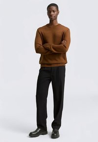 Pruun kudumiga kampsun soonikkoes varrukate ja äärega, mustade pükste ja mustade pahkluu saabastega, käed rüpes seisev.